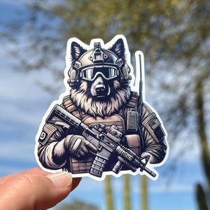 May include: A sticker featuring a wolf in military gear, including a helmet, goggles, and a tactical vest. The wolf is holding a rifle. The sticker has a white border and is held by a hand.