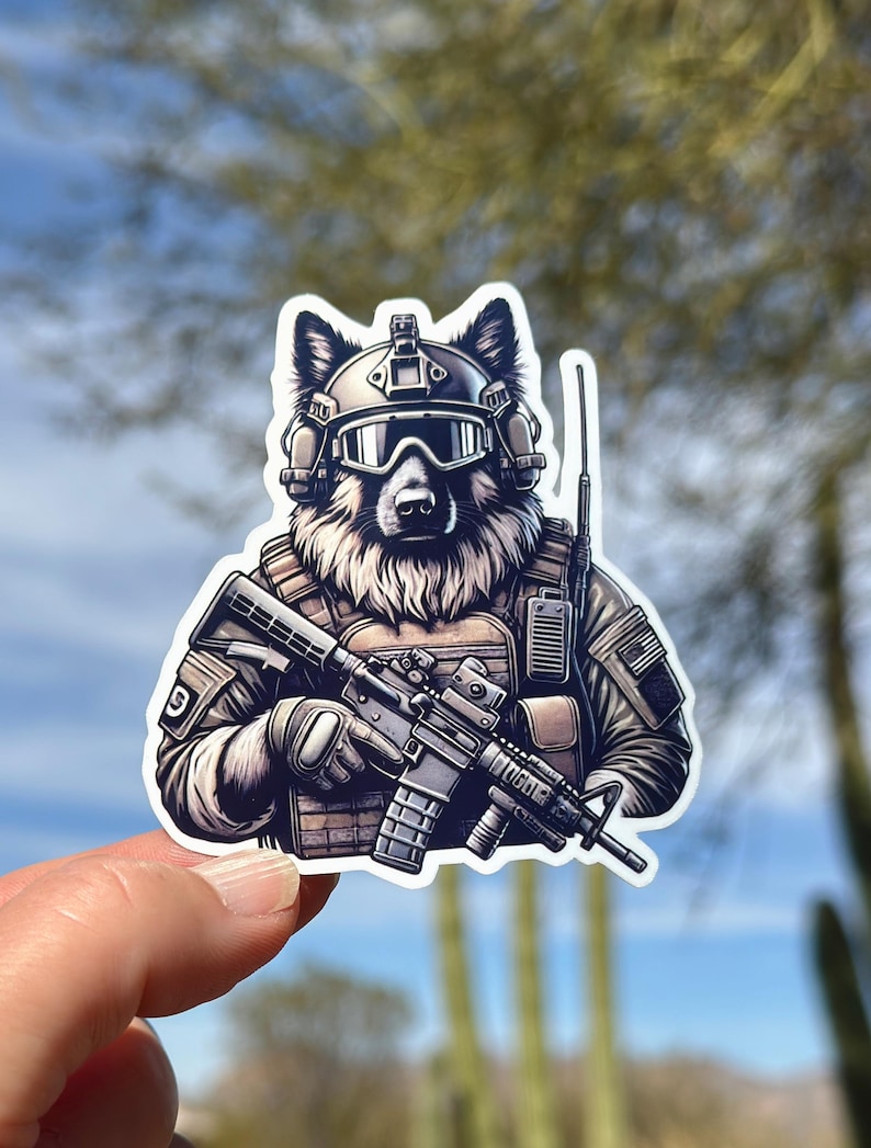 May include: A sticker featuring a wolf in military gear, including a helmet, goggles, and a tactical vest. The wolf is holding a rifle. The sticker has a white border and is held by a hand.