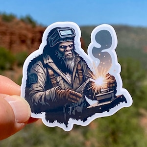 May include: A sticker featuring a cartoon illustration of a furry, brown, ape-like creature wearing a welding mask and welding gear. The creature is welding a metal object with sparks flying from the welding torch.