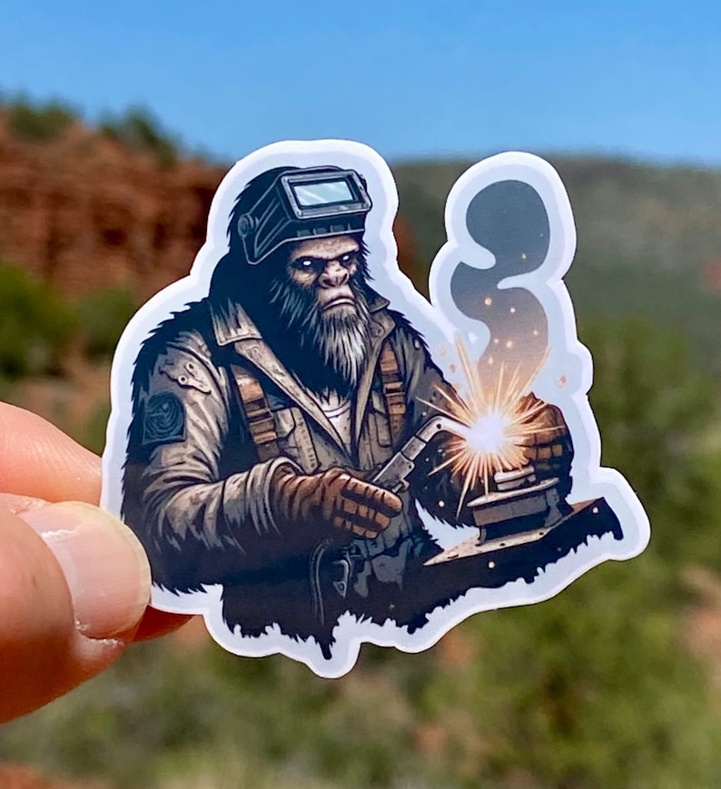 May include: A sticker featuring a cartoon illustration of a furry, brown, ape-like creature wearing a welding mask and welding gear. The creature is welding a metal object with sparks flying from the welding torch.