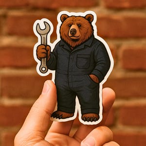 May include: A sticker featuring a cartoon bear in a dark blue mechanic's jumpsuit, holding a silver wrench. The bear has brown fur and is smiling. The sticker is held in front of a brick wall.