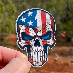May include: A sticker of a skull with the American flag design. The skull has a distressed look with faded red, white, and blue colors.