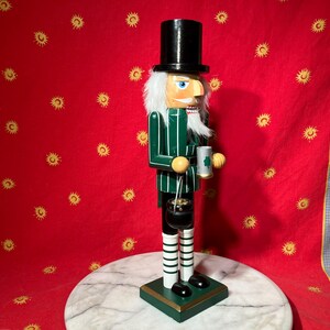 Peut inclure: Un casse-noisette en bois avec une veste rayée verte et blanche, un haut-de-forme noir et une barbe blanche. Le casse-noisette tient une chope avec un trèfle et un pot d'or. La figurine repose sur une base verte.
