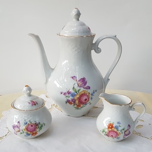 May include: A white porcelain tea set with a floral design. The set includes a teapot, creamer, and sugar bowl. The flowers are pink, yellow, purple, and blue.