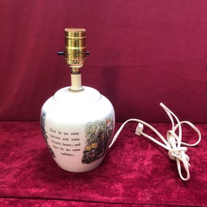 Peut inclure: Lampe de table en céramique blanche avec un support de lampe de couleur laiton et un cordon blanc. La lampe présente une illustration peinte et le texte : "First he ate some lettuces and some French beans; and then he ate some radishes."