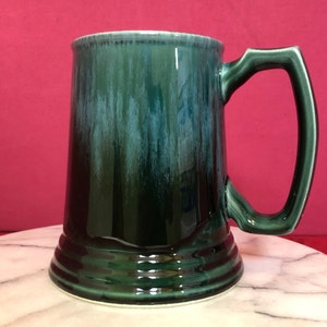 May include: A green ceramic beer stein with a glazed finish. The stein has a wide base and a handle.