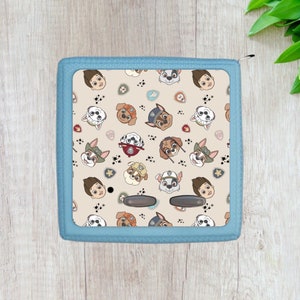 May include: A blue zippered pouch with a beige fabric lining featuring a repeating pattern of cartoon characters from the children's television show Paw Patrol. The characters include Chase, Marshall, Skye, Rocky, Zuma, and Rubble. The pouch has two black buttons on the front.
