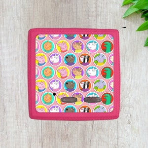 May include: A pink rectangular shaped plush toy with a repeating pattern of cartoon characters in colorful circles. The characters include pigs, a zebra, a dog, an elephant, and a green dinosaur.