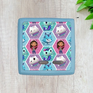 May include: A blue square-shaped cushion with a repeating pattern of pink, purple, and teal hexagons. Each hexagon features a cartoon character, including a girl with brown hair, a blue cat, and a white cat with a pink bow. The cushion has a blue border.