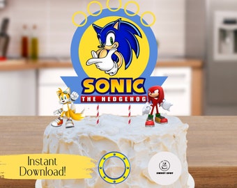 Pode incluir: Um bolo branco com cobertura azul e um topo de bolo do Sonic the Hedgehog. O topo apresenta Sonic, Tails e Knuckles. O bolo é decorado com velas listradas vermelhas e brancas e um círculo amarelo com estrelas.