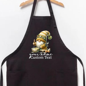 May include: Black apron with a gnome chef design. The gnome is wearing a yellow and white striped hat and holding a bowl and spoon. The text "your name Custom Text" is below the gnome.