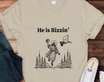 May include: Beige t-shirt with the text "He is Rizzin'" above a black graphic of Jesus dunking a basketball. The graphic includes a basketball hoop and trees.