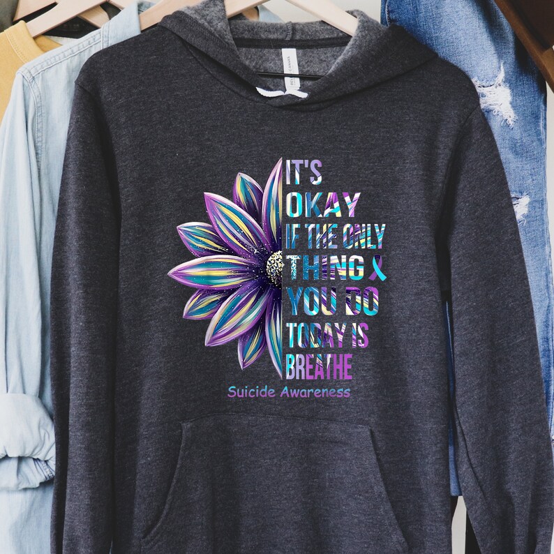 K&ouml;nnte beinhalten: Ein dunkelgrauer Kapuzenpullover mit einem lila, blauen und gelben Blumenmotiv. Der Text "It's okay if the only thing you do today is breathe" ist auf dem Pullover aufgedruckt. Unter dem Text steht der Satz "Suicide Awareness".