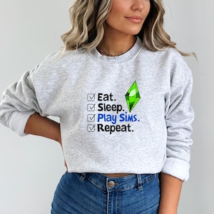 May include: A light gray sweatshirt with the text "Eat. Sleep. Play SIMS. Repeat." in black and blue, with a green diamond graphic. The sweatshirt is a crew neck style.