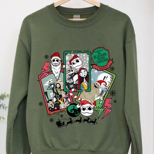 May include: Olive green sweatshirt featuring a graphic design from "The Nightmare Before Christmas." The design includes characters like Jack Skellington, Sally, and others in festive holiday frames, with snowflakes and lightning bolts.