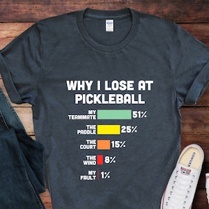 May include: Dark gray t-shirt with the text "WHY I LOSE AT PICKLEBALL" and a bar graph showing the reasons for losing. The reasons include "My Teammate," "The Paddle," "The Court," "The Wind," and "My Fault."