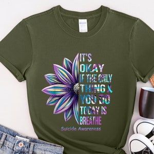 K&ouml;nnte beinhalten: Ein gr&uuml;nes T-Shirt mit einem lila und blauen Blumenmotiv. Der Text auf dem Shirt lautet "It's okay if the only thing you do today is breathe. Suicide Awareness."