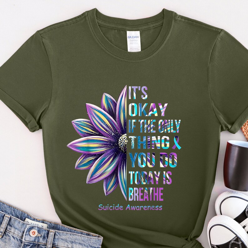 K&ouml;nnte beinhalten: Ein gr&uuml;nes T-Shirt mit einem lila und blauen Blumenmotiv. Der Text auf dem Shirt lautet "It's okay if the only thing you do today is breathe. Suicide Awareness."