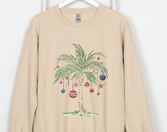May include: A beige sweatshirt featuring a whimsical illustration of a palm tree decorated with colorful ornaments. The tree is drawn in a watercolor style, with green and yellow leaves and various striped and dotted ornaments hanging from the branches.