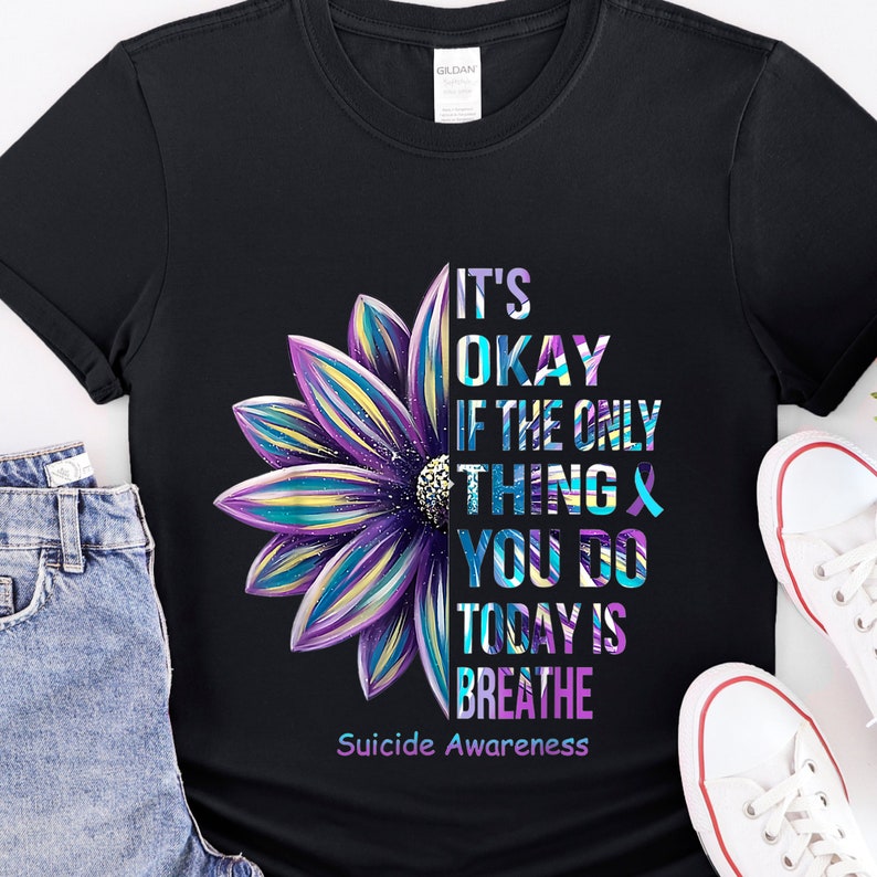 K&ouml;nnte beinhalten: Schwarzes T-Shirt mit einem lila, blauen und gelben Blumenmotiv. Der Text "It's okay if the only thing you do today is breathe" ist auf dem Shirt aufgedruckt. Der Text "Suicide Awareness" ist unter dem Motiv aufgedruckt.
