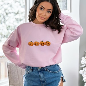 May include: A light pink long-sleeve sweatshirt featuring a horizontal row of four orange jack-o'-lantern pumpkins with distinct carved facial expressions printed across the center of the chest, paired with blue denim shorts.