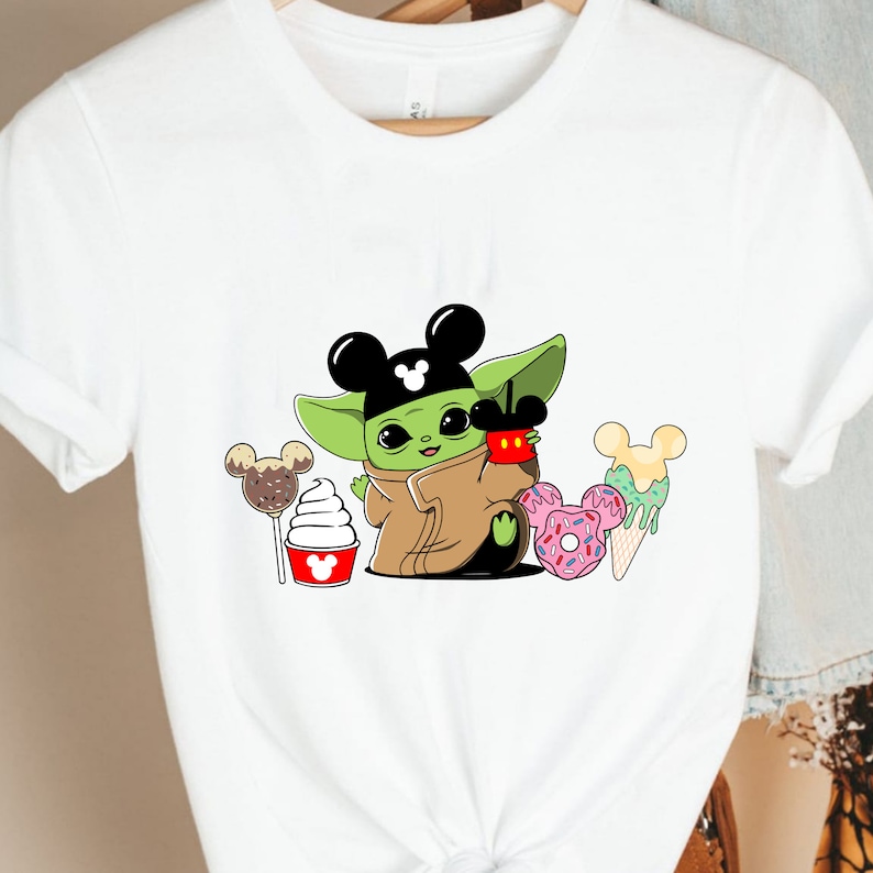 May include: White t-shirt featuring a cartoon character with large ears, wearing a Mickey Mouse hat, holding a red cup. The design includes Mickey Mouse-shaped desserts like ice cream, donuts, and cake pops. The shirt is made of a soft material.