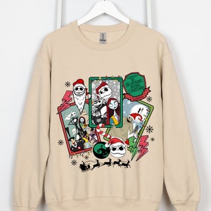 May include: Beige sweatshirt featuring a collage of characters from "The Nightmare Before Christmas" movie. The design includes Jack Skellington in a Santa hat, Sally, and other characters, with snowflakes and text. The sweatshirt is hanging on a white hanger.