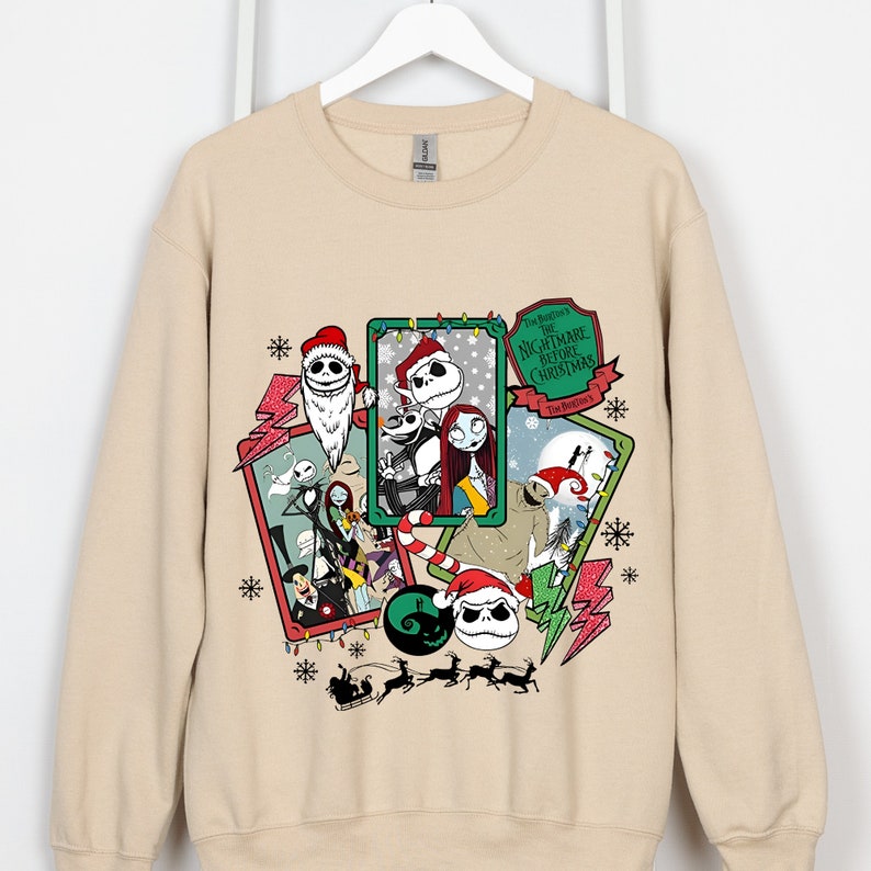 May include: Beige sweatshirt featuring a collage of characters from "The Nightmare Before Christmas" movie. The design includes Jack Skellington in a Santa hat, Sally, and other characters, with snowflakes and text. The sweatshirt is hanging on a white hanger.