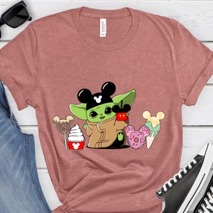 May include: A dusty rose t-shirt featuring a cartoon character with Mickey Mouse ears, holding a cup and surrounded by desserts. The character is green with a brown cloak. Desserts include a lollipop, ice cream, and donuts.