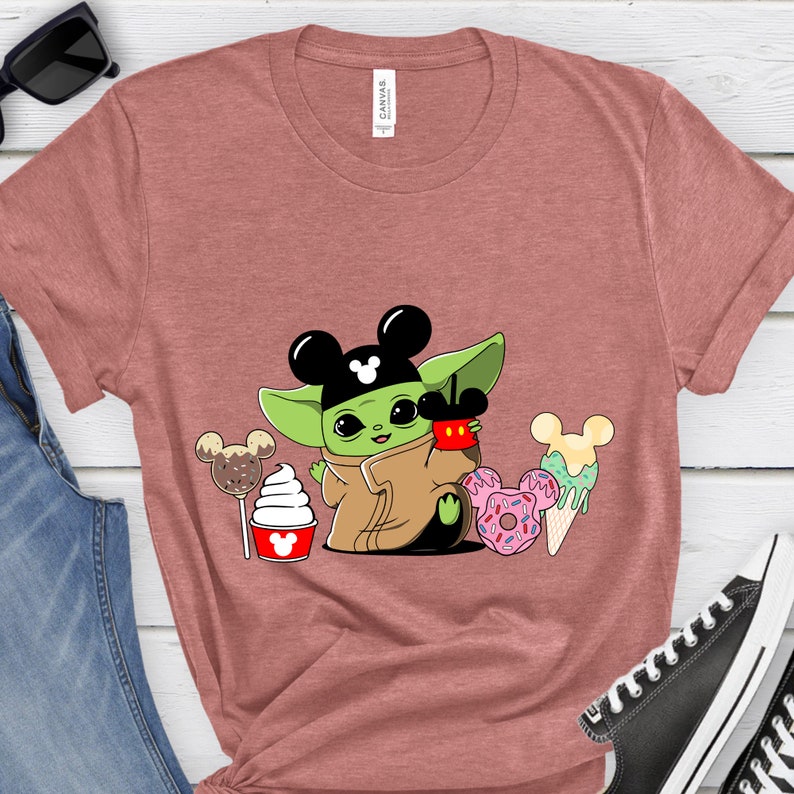 May include: A dusty rose t-shirt featuring a cartoon character with Mickey Mouse ears, holding a cup and surrounded by desserts. The character is green with a brown cloak. Desserts include a lollipop, ice cream, and donuts.