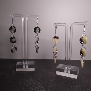 May include: Two pairs of dangle earrings with a silver chain and three round buttons. One pair has black and white buttons, the other pair has cream and brown buttons.