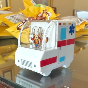 May include: A white ambulance-shaped keychain featuring a clear plastic cab with a cartoon giraffe character wearing a medical head mirror and blue face mask inside. The vehicle has a red stripe, blue windows, and a blue Star of Life emblem.