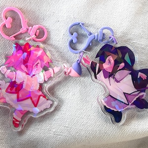 May include: Two iridescent acrylic charms featuring anime characters. The charm on the left is pink and features a girl with pink hair and a pink dress. The charm on the right is purple and features a girl with dark purple hair and a white dress.