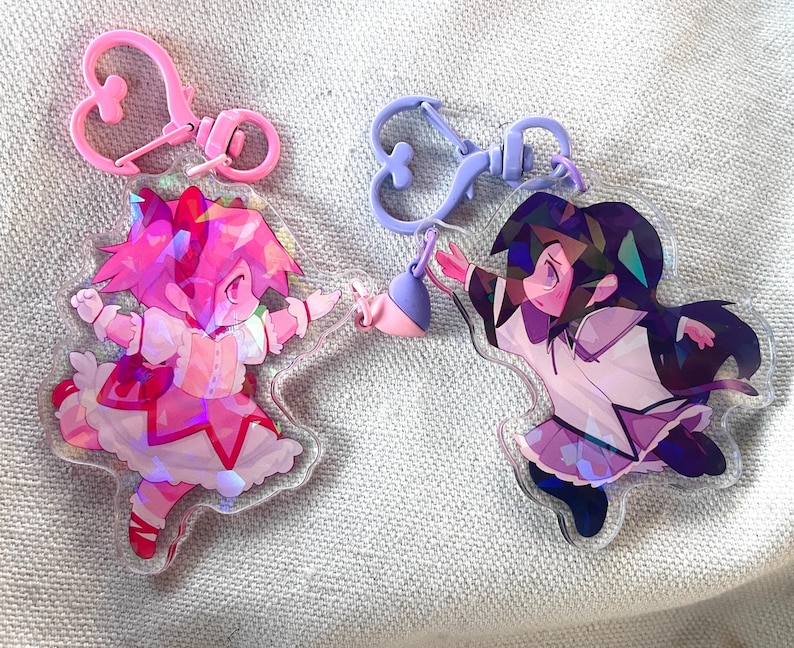May include: Two iridescent acrylic charms featuring anime characters. The charm on the left is pink and features a girl with pink hair and a pink dress. The charm on the right is purple and features a girl with dark purple hair and a white dress.