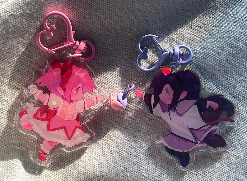 May include: Two anime character keychains, one pink and one purple, with heart-shaped keyrings. The pink character is wearing a pink and white dress with a star pattern. The purple character is wearing a white and purple dress with a star pattern.