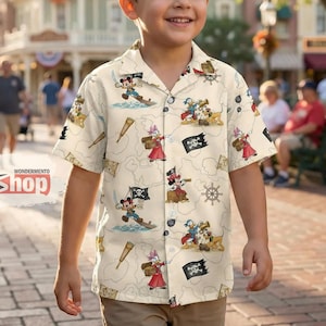 May include: A short-sleeved, button-down shirt with a pirate-themed pattern featuring cartoon characters. The shirt is cream-colored with illustrations of pirates, treasure maps, and nautical elements. The shirt is worn by a young boy.