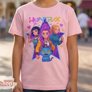 May include: Pink t-shirt featuring a graphic of three female characters and a blue cat, set against a star-shaped background. The word "HUNTR/X" is printed above the characters. The shirt is worn by a person.