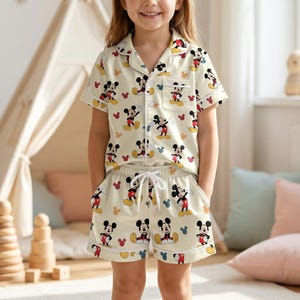 K&ouml;nnte beinhalten: Ein Kind tr&auml;gt einen cremefarbenen Pyjama mit einem sich wiederholenden Mickey Mouse-Muster. Das kurz&auml;rmelige Oberteil hat eine Tasche und Kn&ouml;pfe, und die Shorts haben einen Kordelzug in der Taille. Das Pyjama-Set ist rot und wei&szlig; eingefasst.