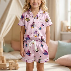 May include: Pink pajama set featuring a repeating pattern of animated characters. The short-sleeved top and shorts have white trim and a drawstring waist. The child is wearing the pajamas and smiling at the camera.