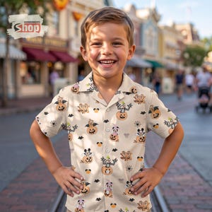 May include: A short-sleeved button-down shirt in a light beige color featuring an all-over print of various cartoon characters dressed in Halloween costumes, sitting inside orange jack-o'-lantern pumpkins, along with small black bats and spider webs.