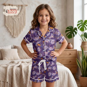 May include: Purple pajama set with a whimsical print of cartoon characters, featuring a button-down short-sleeve top and matching shorts. The set has white trim and a drawstring waist. The Wondermento Shop logo is visible in the upper left corner.