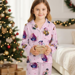 May include: A light pink pajama set with a repeating pattern of animated characters and musical notes. The long-sleeved top and pants are button-down and feature a collared neckline. The pajama set is worn by a young person holding a small gift.
