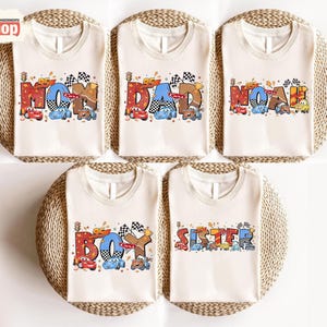 May include: Five off-white t-shirts with racing-themed designs. Each shirt features a different word: "MOM", "DAD", "NOAH", "BOY", and "SISTER", with colorful race cars and checkered flags. The text is in various colors and fonts.