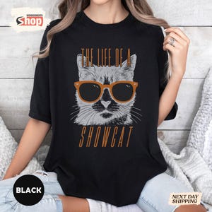 May include: Black t-shirt featuring a graphic of a cat wearing sunglasses. The cat is gray and white, with orange-framed sunglasses. The text "THE LIFE OF A SHOWCAT" is printed above and below the cat image in orange.