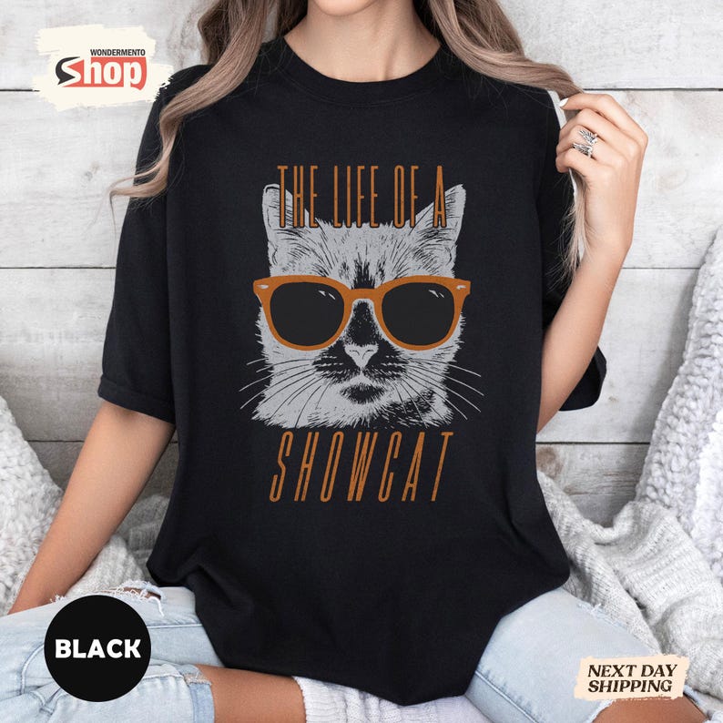 May include: Black t-shirt featuring a graphic of a cat wearing sunglasses. The cat is gray and white, with orange-framed sunglasses. The text "THE LIFE OF A SHOWCAT" is printed above and below the cat image in orange.