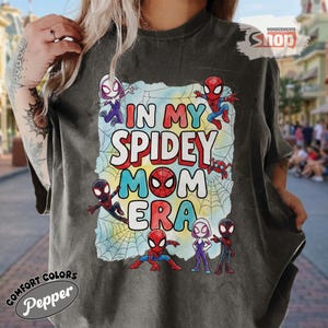 May include: Dark grey t-shirt with the text "IN MY SPIDEY MOM ERA" in colourful letters. The design features Spider-Man characters and a spiderweb background. The shirt has the "Comfort Colors" and "Pepper" logos.