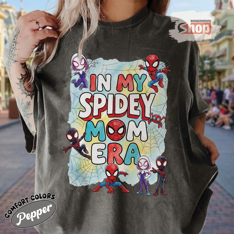May include: Dark grey t-shirt with the text "IN MY SPIDEY MOM ERA" in colourful letters. The design features Spider-Man characters and a spiderweb background. The shirt has the "Comfort Colors" and "Pepper" logos.