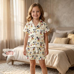 K&ouml;nnte beinhalten: Ein Kind tr&auml;gt einen cremefarbenen Schlafanzug mit einem sich wiederholenden Mickey Mouse-Muster in Rot, Schwarz, Gelb und Blau. Das Schlafanzugoberteil hat kurze &Auml;rmel und eine Knopfleiste. Die Shorts haben einen Kordelzug in der Taille. Das Logo "WONDERMENTO Shop" ist sichtbar.