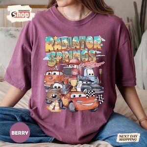 May include: Berry-coloured t-shirt featuring a colourful graphic of characters from the film Cars, including Lightning McQueen and Mater, with the text "Radiator Springs". The design includes a chequered flag and a trophy.