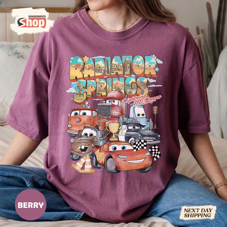 May include: Berry-coloured t-shirt featuring a colourful graphic of characters from the film Cars, including Lightning McQueen and Mater, with the text "Radiator Springs". The design includes a chequered flag and a trophy.